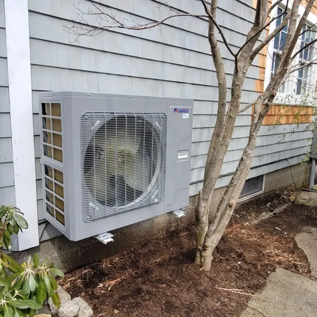 A large air conditioner is mounted on the side of a house next to a tree.