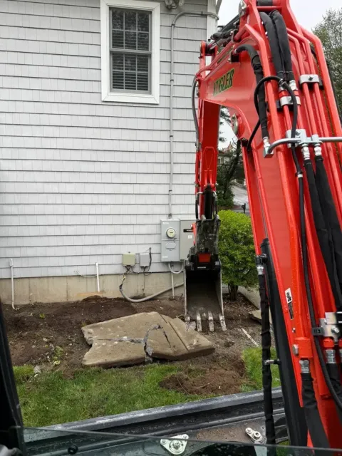 Red excavator bucket beside a house foundation with a concrete slab and exposed utility lines