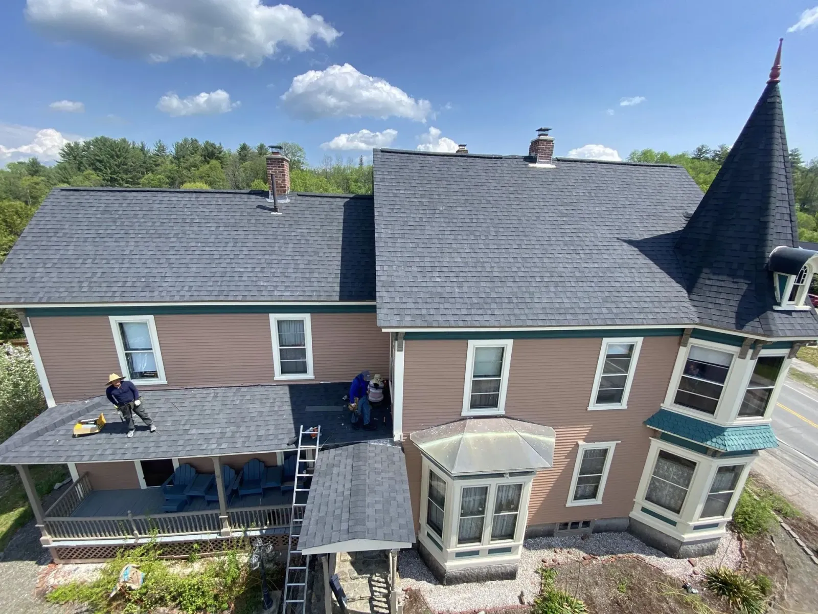 Aerial view of a house with gray roof, tan siding, and a covered porch, with workers on the roof.