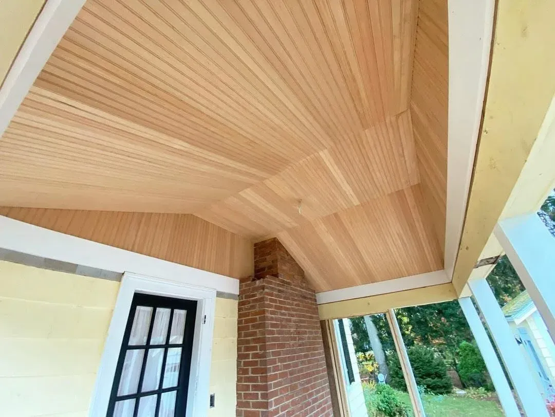 View of a porch ceiling with wooden panels, brick chimney, and a black window with white trim, part of a yellow house with white trim surrounded by green trees.
