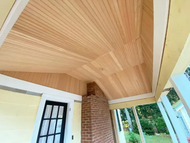 View of a porch ceiling with wooden panels, brick chimney, and a black window with white trim, part of a yellow house with white trim surrounded by green trees.