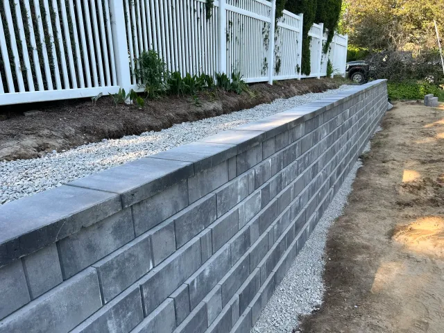 Construction of a gray stone retaining wall with gravel and soil behind, white metal fence on top, and green plants along the fence in a yard.