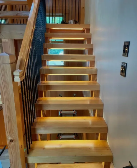 Unfinished wooden staircase with treads, railing and a black spindled guard on the left side, against a cream-colored wall with two electrical outlets on the right, during construction.