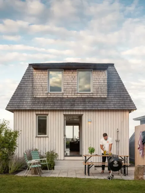 A man grills food on a barbecue in front of a small two-story wooden house, crafted by Maine homebuilders, under a partly cloudy sky.