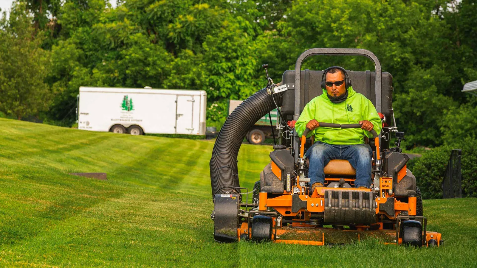 Man in green hoodie and sunglasses operating an orange lawn mower on grass near trees.