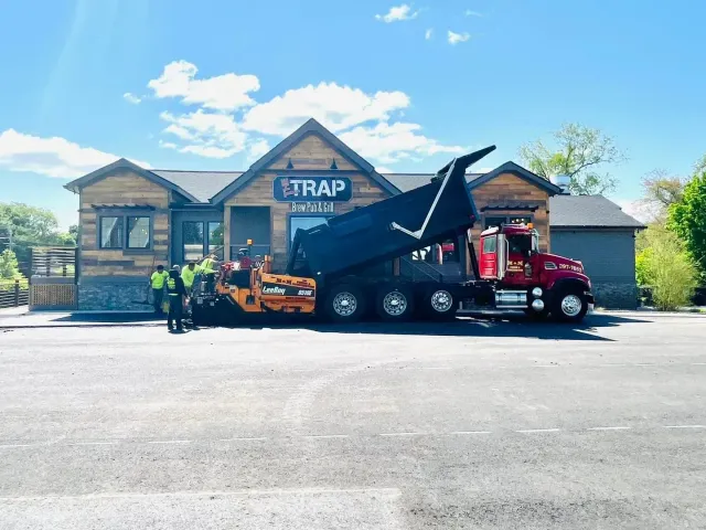 Asphalt paving at TRAP Bar & Grill. Dump truck unloading asphalt into a paving machine. Two workers stand near the machine.