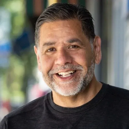 Smiling man with salt-and-pepper beard in a black shirt, standing outdoors in soft daylight.