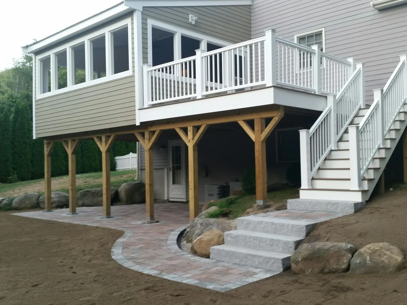 A house with a raised deck and staircase, under construction, with wooden support beams and a brick patio.