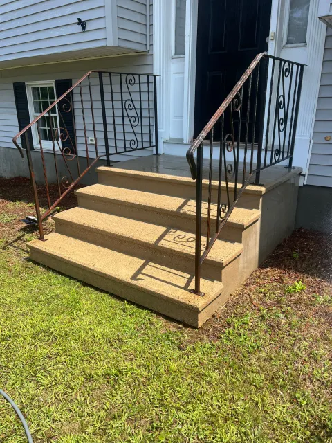 Front porch steps with tan concrete stairs and black railings beside a house, bordered by grass.