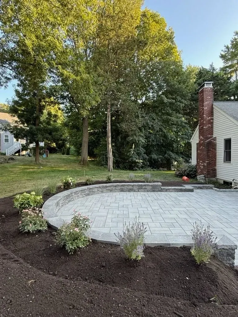 A curved stone patio with a low retaining wall, surrounded by fresh mulch and young shrubs, next to a brick-chimney house.