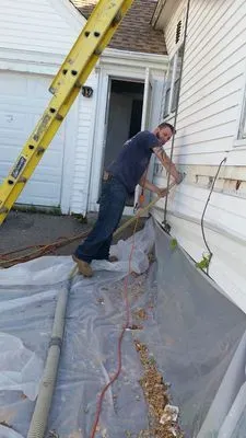 A man works on the side of a white house. He is standing on a plastic sheet. A ladder is propped against the house.