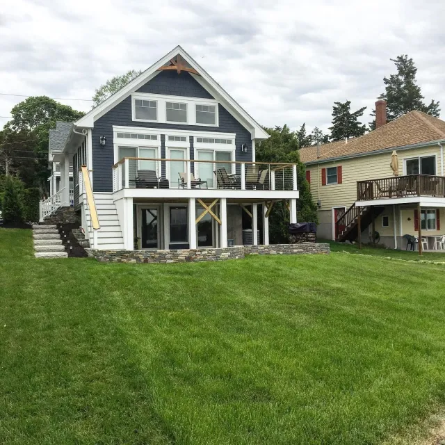 A two-story blue house with a white-railed deck and stone patio, situated next to a yellow house with a brown roof.