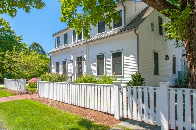 A white two-story house with black shutters, a gabled roof, and a white picket fence is surrounded by green grass, trees, and a brick walkway under a clear blue sky.
