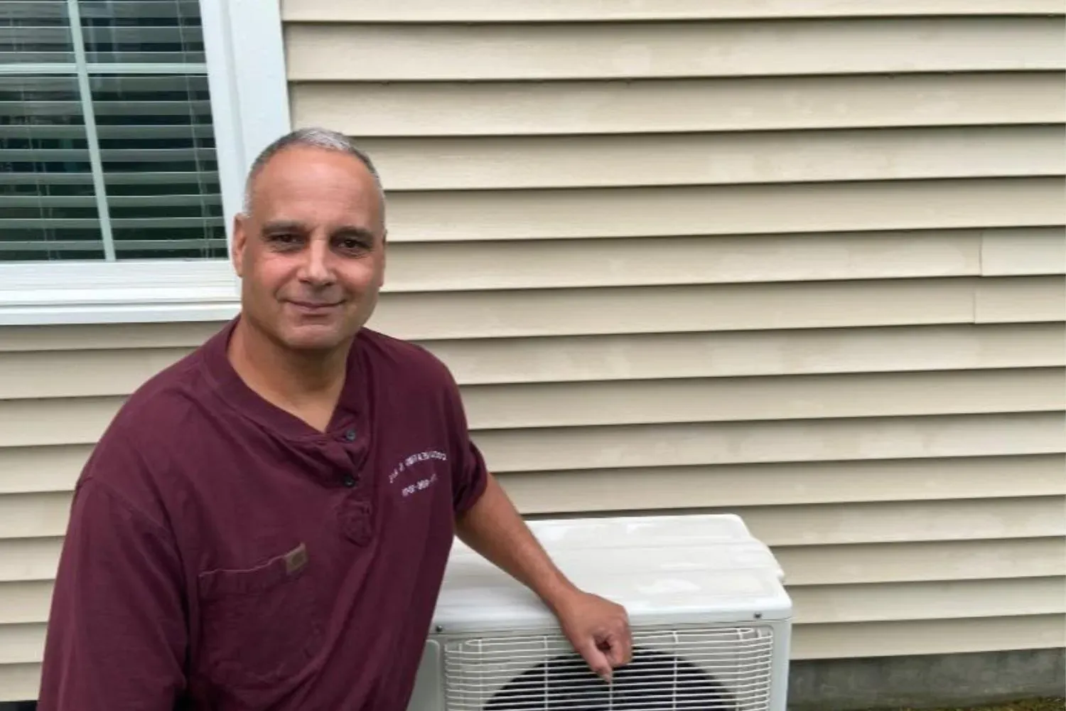 Man in maroon shirt standing beside an outdoor air conditioner unit next to a house.
