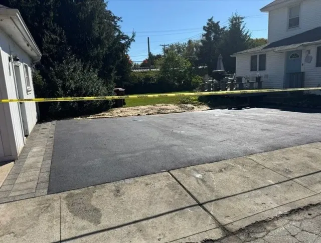 Newly paved asphalt driveway with brick border next to a white garage and house. Yellow caution tape.