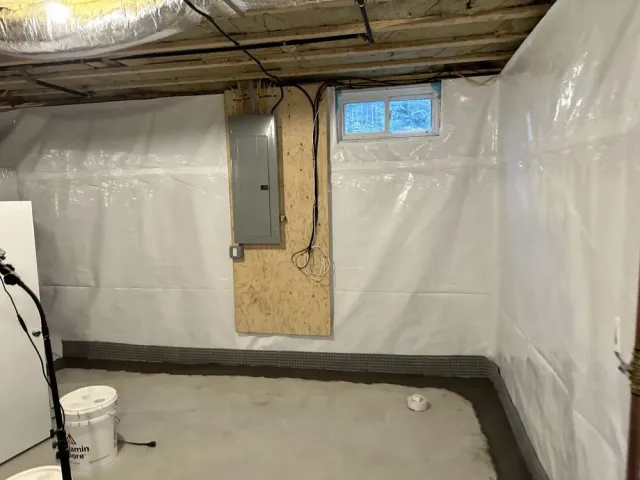 Unfinished basement with plastic-covered walls, exposed ceiling, electrical panel, and small window.