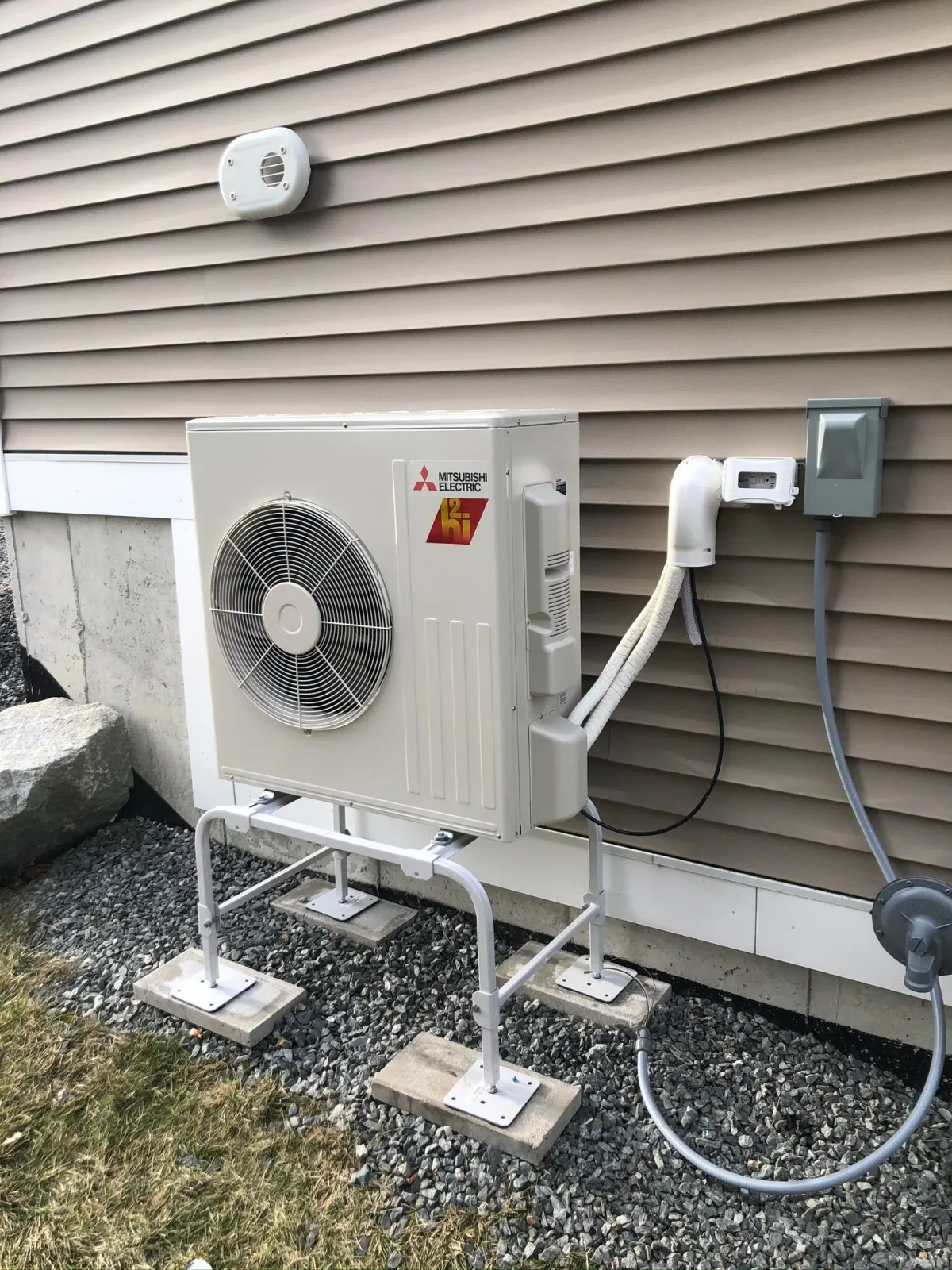 Outdoor Mitsubishi Electric heat pump unit mounted on a stand next to a building with beige siding.