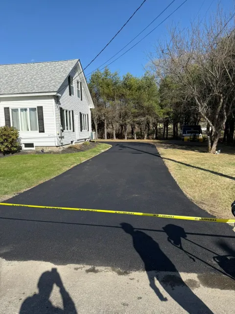 A newly paved black asphalt driveway leads to a white house with a yellow caution tape stretched across the entrance.