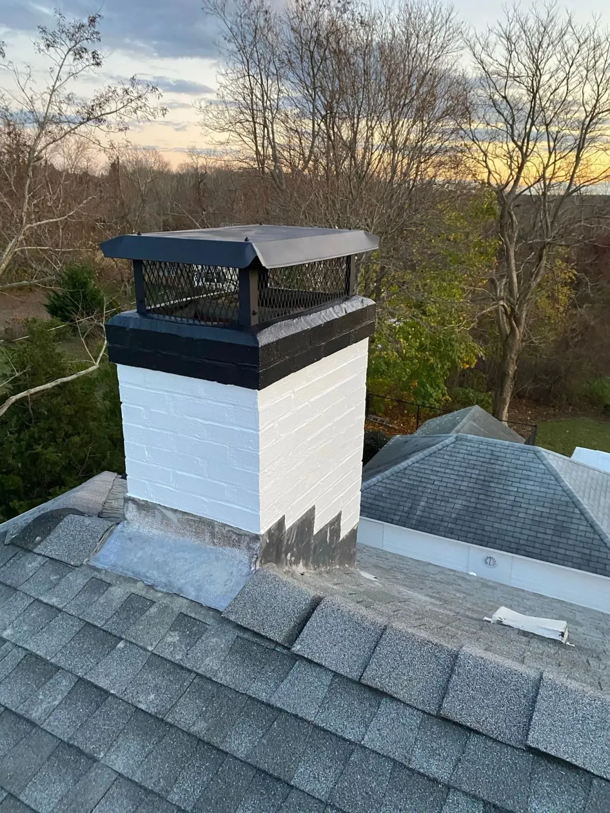A white painted brick chimney with a black metal cap, situated on a shingled roof against a backdrop of trees at dusk.