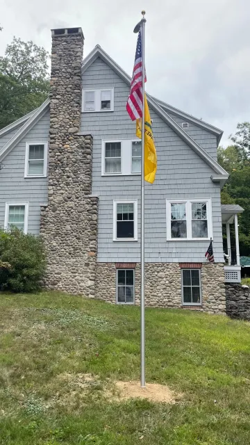 A house with a large stone chimney, gray siding, and white-framed windows. An American flag, a yellow flag, and a small American flag are flying on a single flagpole in front of the house.