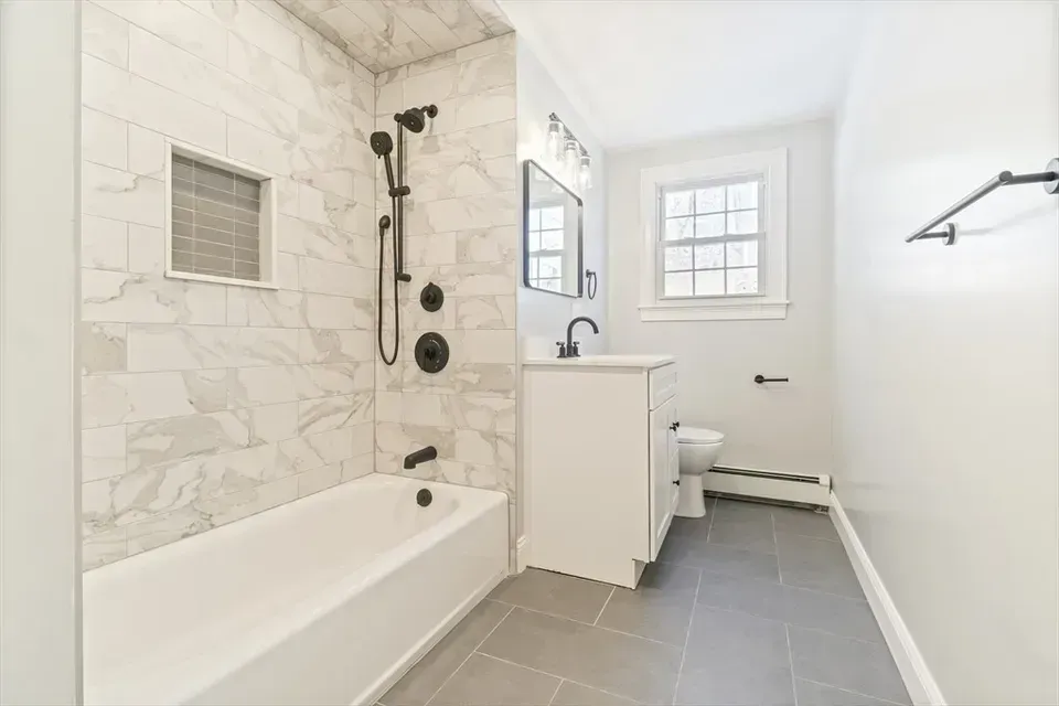 A bathroom with a white tub, marble-tiled shower, white vanity, and gray floor tiles under a window.