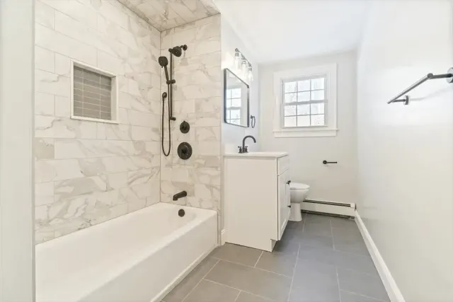 A bathroom with a white tub, marble-tiled shower, white vanity, and gray floor tiles under a window.