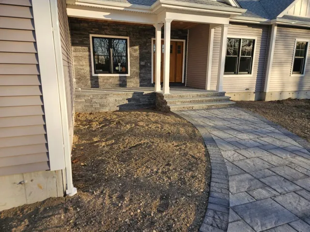 A house exterior with a paved walkway leading to the front steps. Brown siding and stone facade visible.