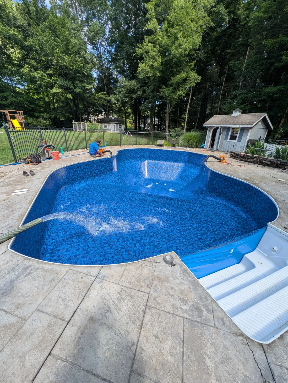 A backyard with a kidney-shaped in-ground swimming pool, one person working on the pool, a small shed, a fenced yard, a play set, and a surrounding forested area.