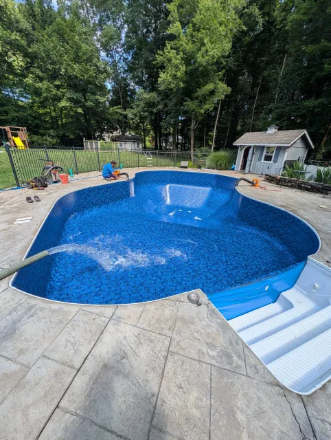 A backyard with a kidney-shaped in-ground swimming pool, one person working on the pool, a small shed, a fenced yard, a play set, and a surrounding forested area.