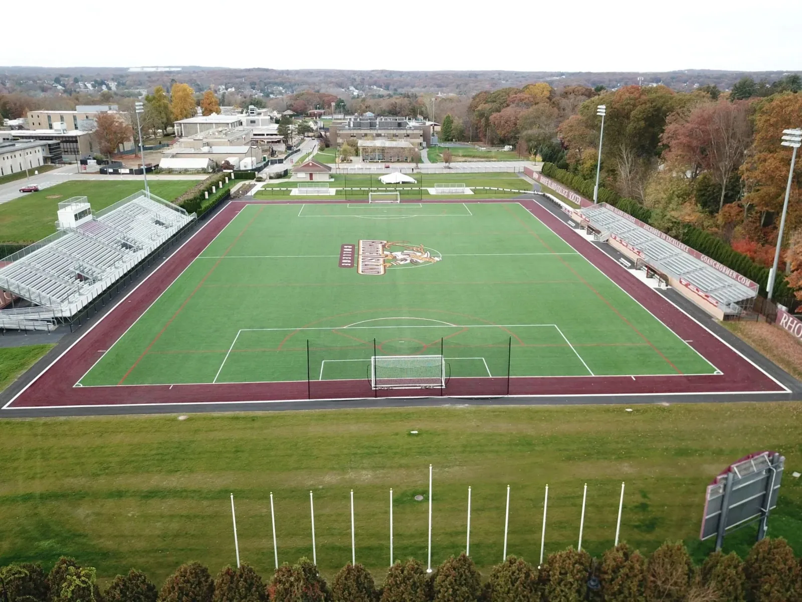 An aerial view of an outdoor sports field with a green soccer field and surrounding track, stadium seating on both sides, and trees in the background.