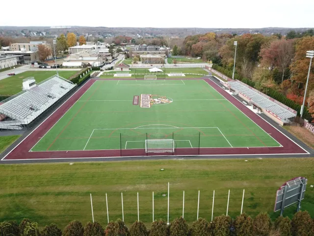 An aerial view of an outdoor sports field with a green soccer field and surrounding track, stadium seating on both sides, and trees in the background.