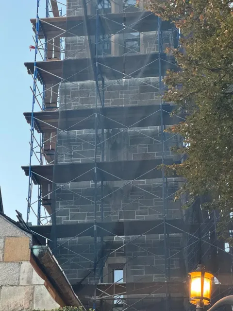 A tall stone tower under renovation, surrounded by blue scaffolding and black construction netting against a bright sky.