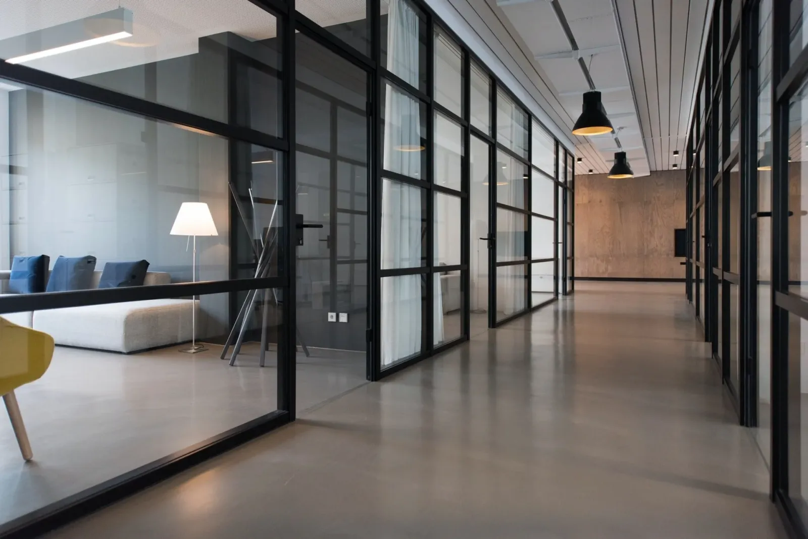 Modern office corridor with glass partitions, black metal framing, and minimalist decor, including a sitting area with a floor lamp visible through the glass.