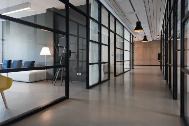 Modern office corridor with glass partitions, black metal framing, and minimalist decor, including a sitting area with a floor lamp visible through the glass.