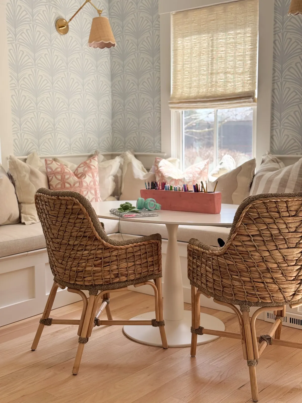 Hardwood floor refinishing in a Marshfield kitchen nook with a white round table, wicker chairs, a built-in bench with beige cushions and pink patterned pillows, a window with a beige shade, and a pink container of colored pencils on the table.