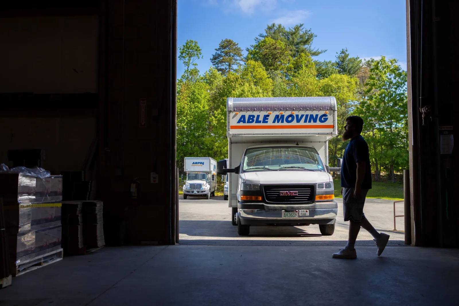 A man with a beard walking past a white moving truck labeled "Able Moving" outside a warehouse or storage unit, with trees and a blue sky in the background.