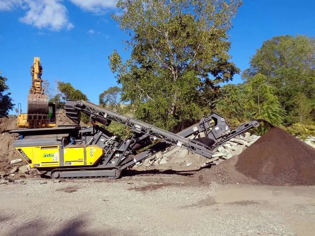 Yellow industrial machinery, excavator, and pile of crushed stone in an outdoor setting under a blue sky.