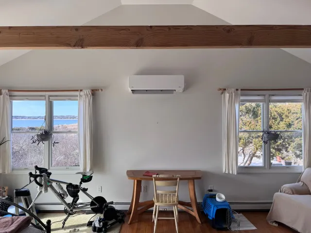 Bright living room with two windows, wall AC, wooden beam, and a bicycle near a small table and couch.