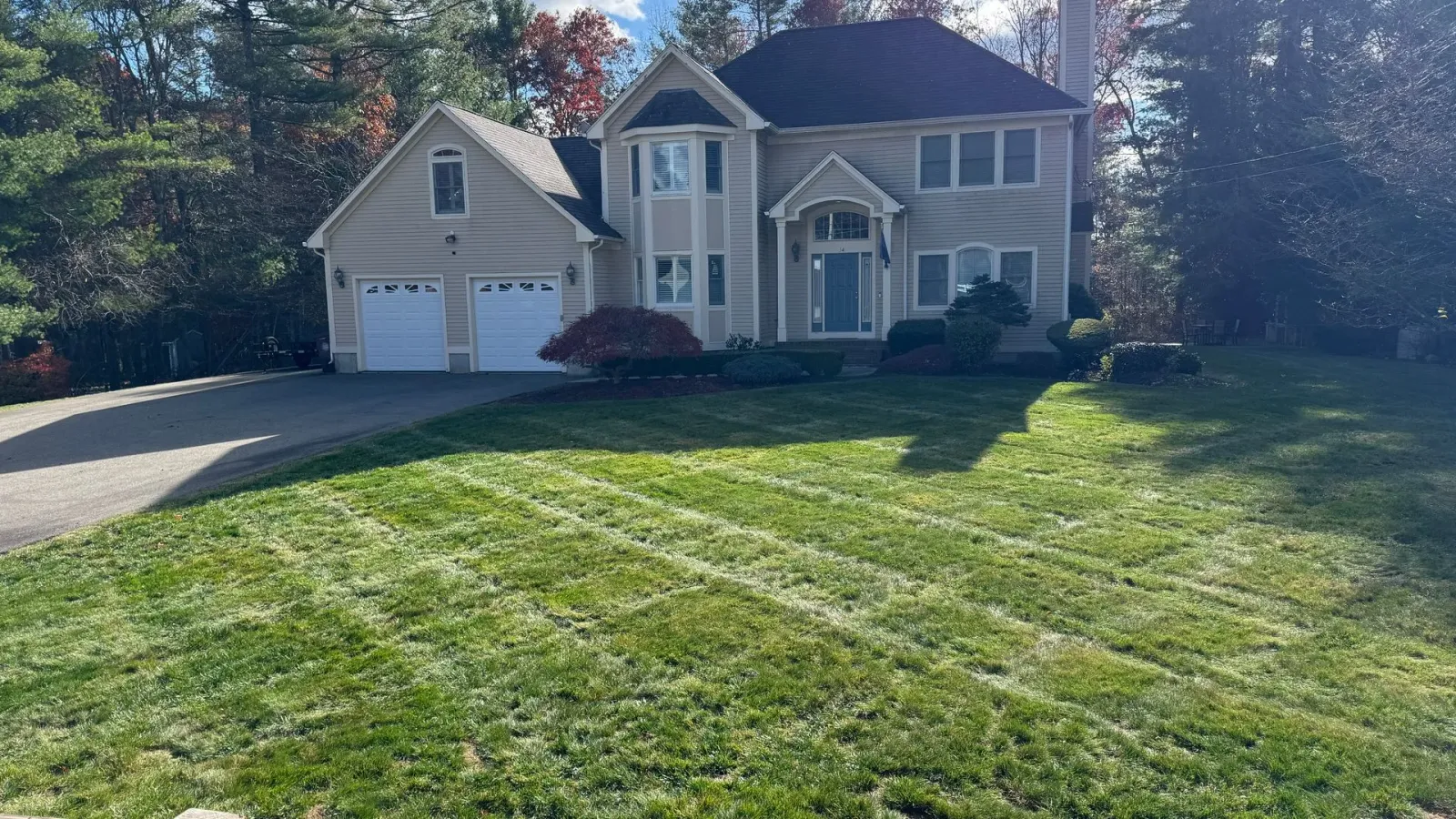 Beige two-story house with attached garage, green lawn, and trees. Blue sky visible.