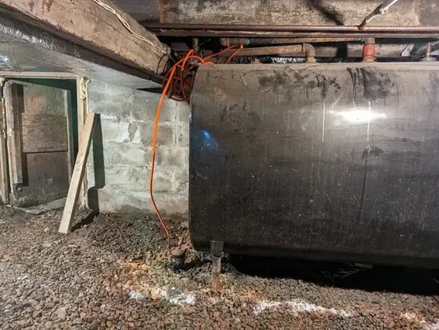 A large, dark cylindrical oil tank sits in a basement with exposed wooden beams, stone walls, and gravel flooring.