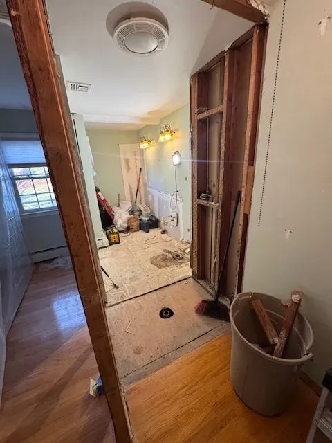 A doorway view of a room under renovation with exposed wooden wall studs, tools, and a trash bin on a wooden floor.