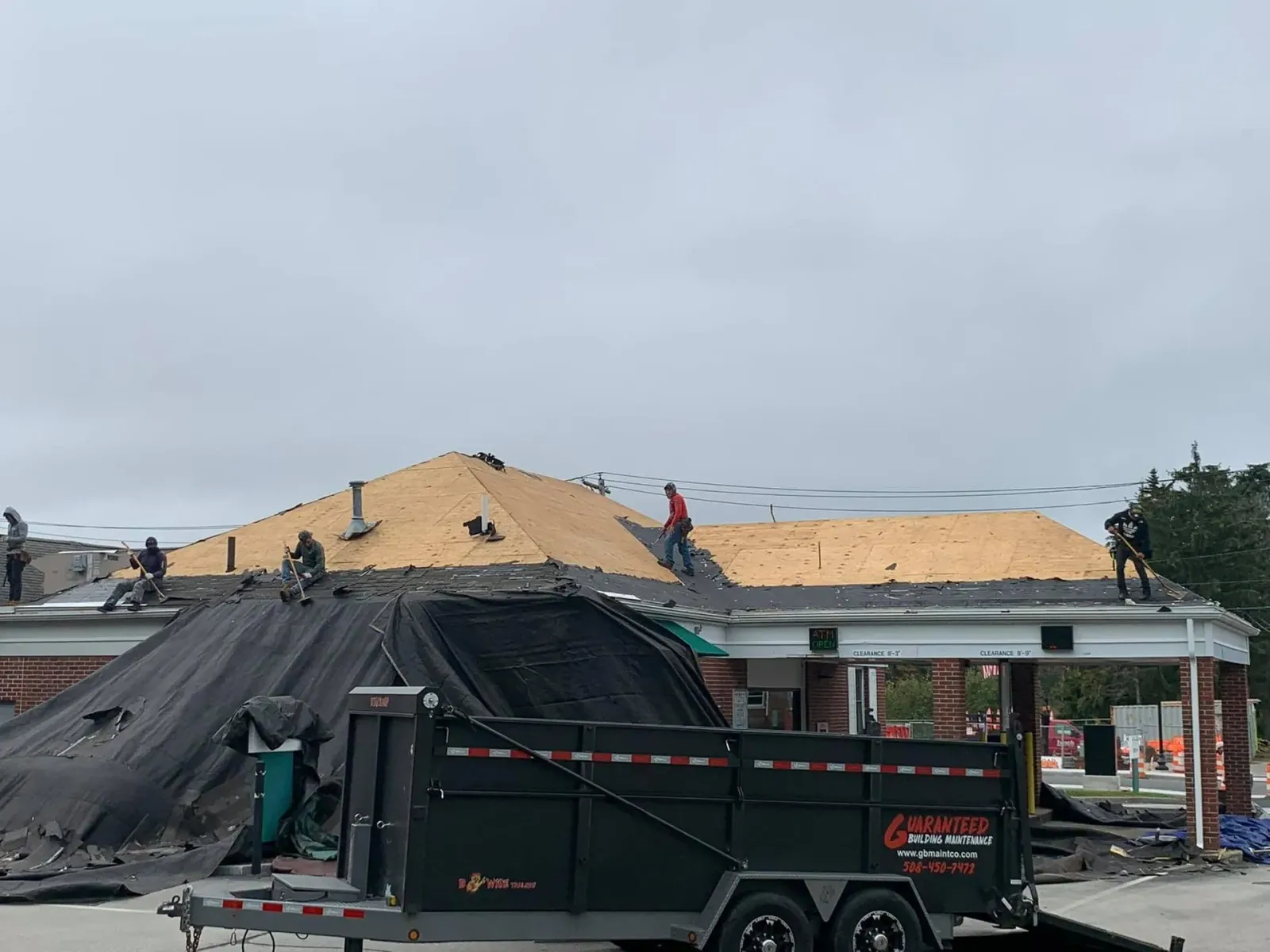 Roofing crew installing plywood on a house under construction, with black tarp and green dumpster in front