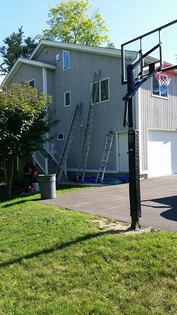 House exterior with ladders leaning against gray siding beside a driveway and basketball hoop