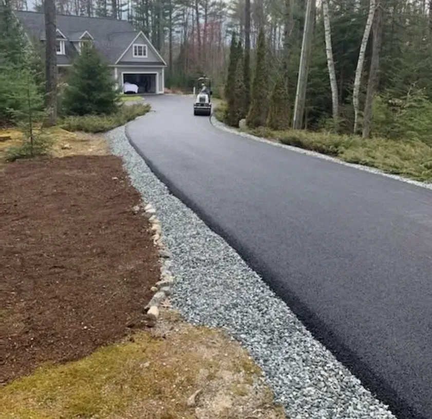 A newly paved asphalt driveway leads to a suburban home, bordered by a strip of gray gravel and a lawn area.