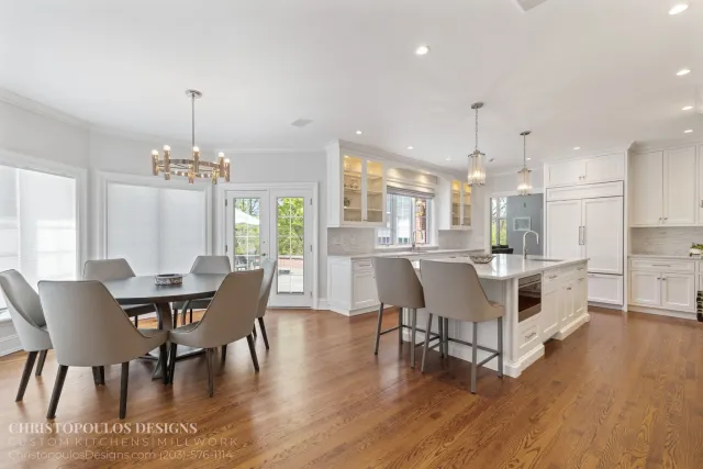 Open-concept kitchen and dining area with white cabinetry, a kitchen island with barstools, a dining table with beige chairs, large windows, and modern lighting fixtures.