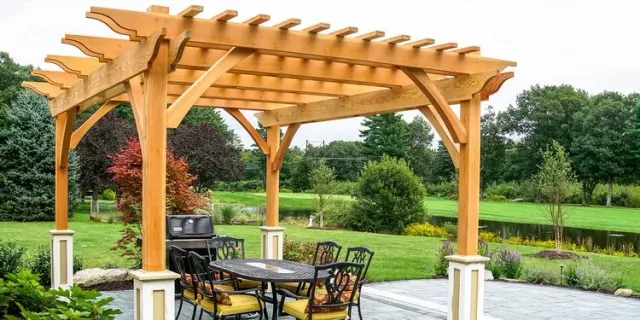 Custom pergola and outdoor dining area installed by Artisan Grounds in Greenland, NH, surrounded by lush landscaping with a natural pond backdrop.