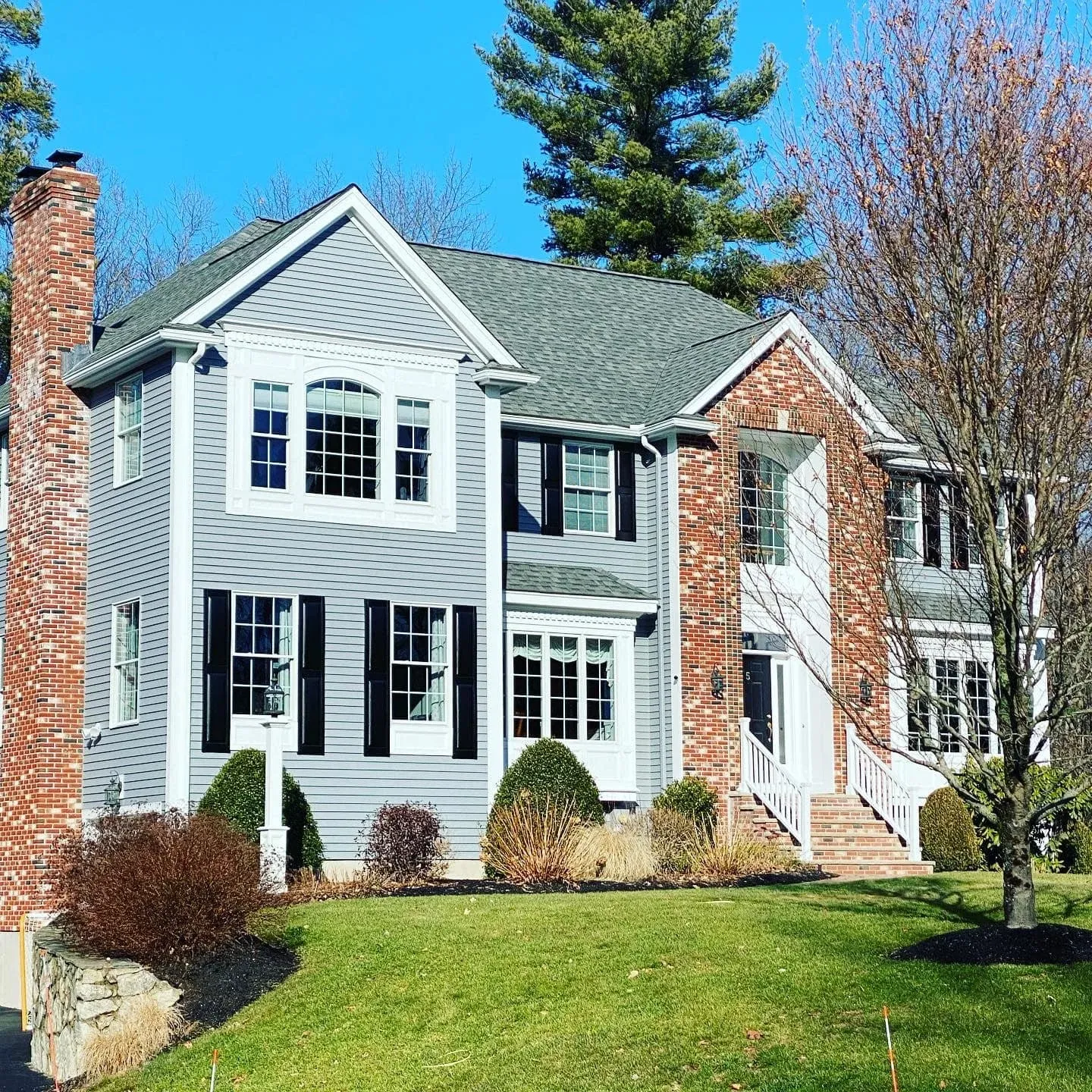 Front view of a two-story suburban house with gray siding, red brick accents, black shutters, white trim, and a green roof, surrounded by a landscaped lawn with bushes and trees under a clear blue sky.