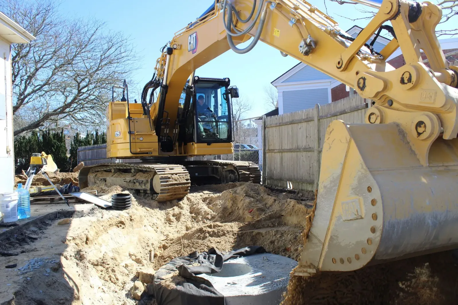 Yellow excavator digging near a wooden fence and building on a sunny day.