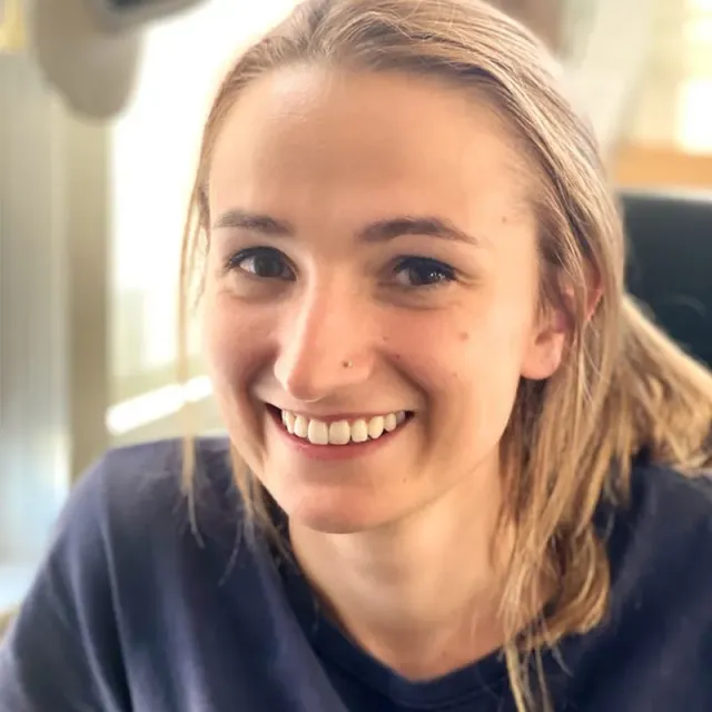 Woman with blonde hair smiles, wearing a navy blue shirt; indoor setting.