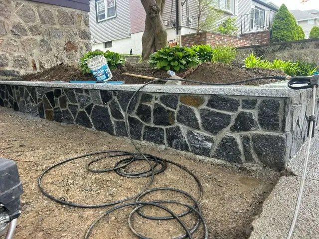 Stone retaining wall with dark irregular stones. Ground-level view. Wires and supplies present.